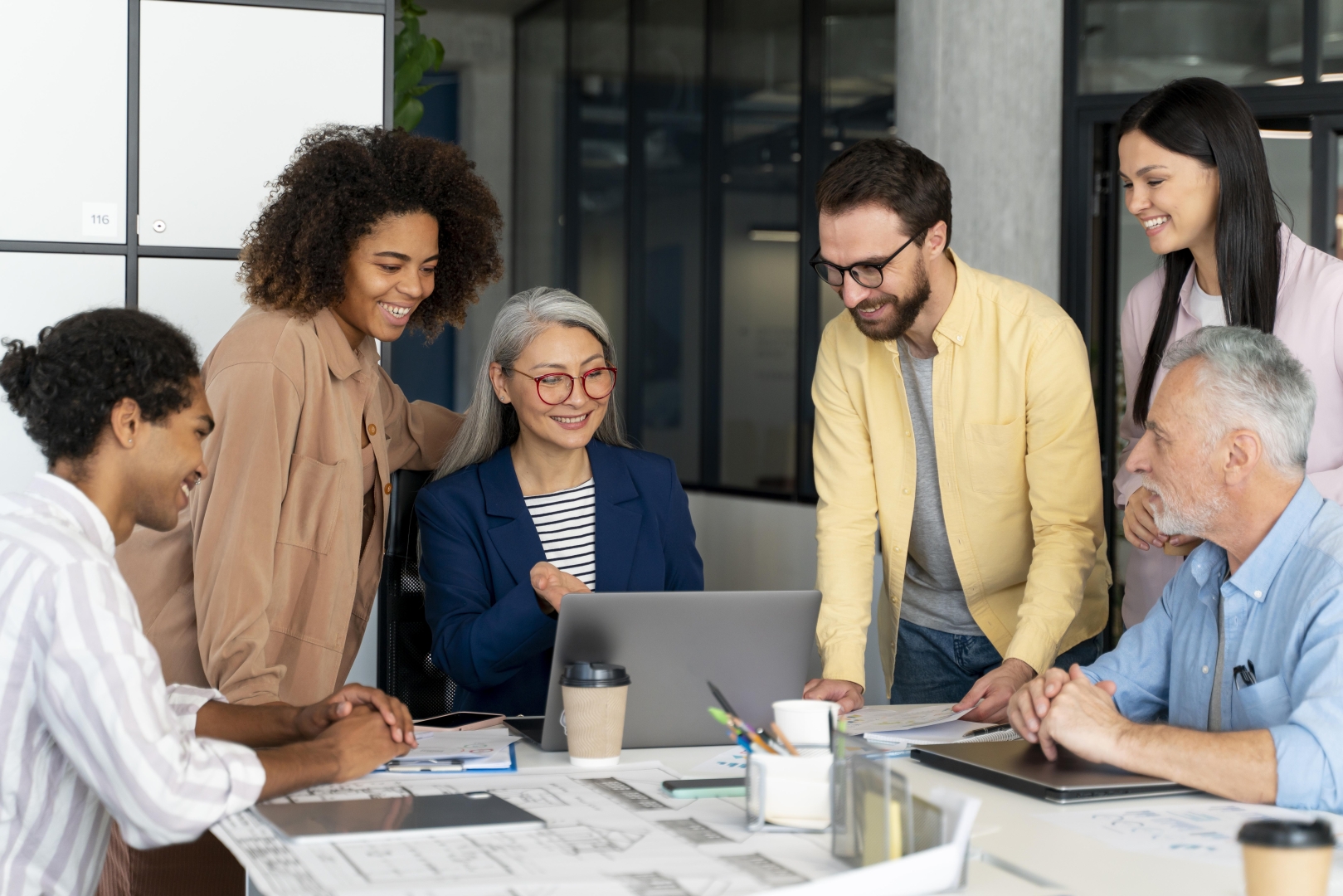 Six coworkers stand around a table with blueprints on it.